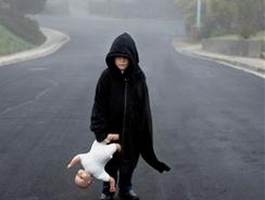 เด็กชายสวมเสื้อคลุมสีดำเดินกลางถนน ในมือถือตุ๊กตาเด็กทารก